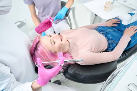 Foto de Paciente en jeans se encuentra en la silla dental, el dentista ha preparado una jeringa con anestesia - Imagen libre de derechos