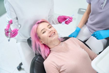 Foto de Mujer sonriente con el pelo rosa y el personal médico se encuentran en el consultorio dental, el médico tiene jeringa y una herramienta especial - Imagen libre de derechos