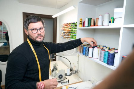Photo for Sad tailor takes a spool from a shelf with a measuring tape hanging around his neck - Royalty Free Image