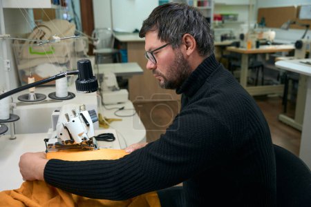Foto de Sastre masculino trabaja en un overlocker, su lugar de trabajo está bien iluminado, un maestro en gafas y un suéter de punto - Imagen libre de derechos