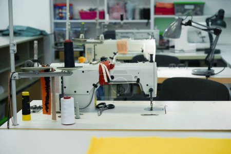 Foto de Máquinas de coser modernas en las mesas del taller, sobre el fondo de estanterías con materiales de costura - Imagen libre de derechos