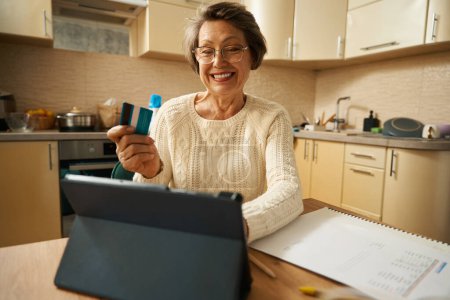 Vieille femme avec des lunettes assis à la table avec tablette, tenant la carte de crédit et regardant le papier