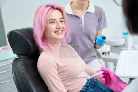 Foto de Mujer sonriente de pelo rosado con frenos en la recepción del dentista, el médico y el asistente utilizan herramientas especiales - Imagen libre de derechos