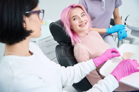 Foto de Una joven sonriente se sienta cómodamente en una silla dental, una ortodoncista la consulta - Imagen libre de derechos