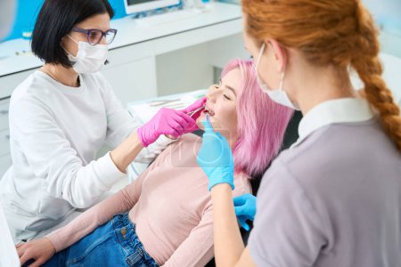 Photo for Orthodontist woman and assistant adjust the bracket system to a young patient, they use an irrigator - Royalty Free Image