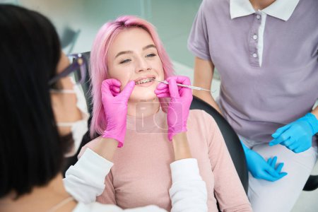 Foto de Joven mujer de pelo rosa en una cita con un ortodoncista, ella está siendo atendida por sus aparatos ortopédicos para alinear sus dientes - Imagen libre de derechos