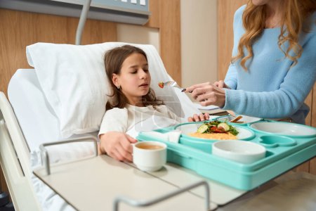 Schwaches Mädchen liegt auf Krankenhausbett, Mutter füttert ihr Krankenhausfutter