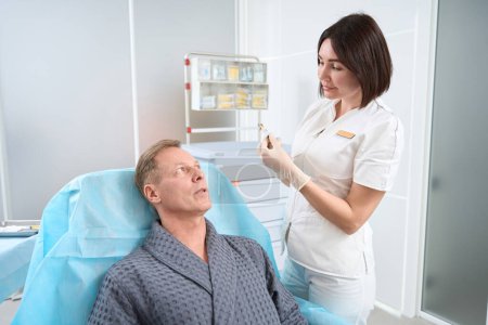 Besorgter männlicher Patient schaut besorgt, während er im Behandlungsstuhl in der Klinik neben einer Ärztin mit Spritze in der Hand sitzt