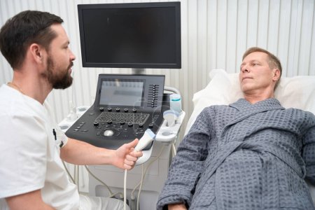 Téléchargez les photos : Skillful medical specilaist talking to a patient before performing an ultrasound check-up of his joints - en image libre de droit