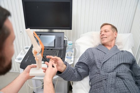 Foto de Male patient on a hospital bed pointing to a replica of human knee in doctors hands - Imagen libre de derechos