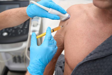 Foto de Cropped photo of doctor in surgical gloves pressing an ultrasound probe to person shoulder and inserting a syringe needle - Imagen libre de derechos
