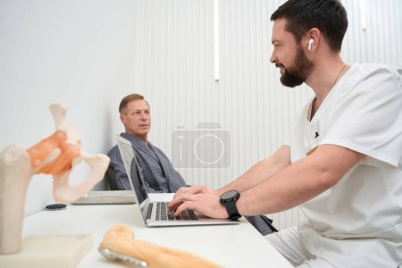 Téléchargez les photos : Certified orthopaedist sitting at clinic desk while having an appointment with a male patient - en image libre de droit