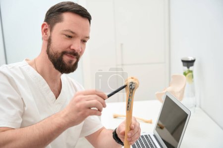 Foto de Friendly medical doctor smiling while holding a replica of human bone and pointing to a part of it with pen - Imagen libre de derechos