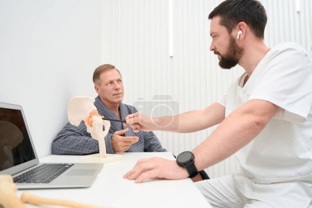 Foto de Professional orthopaedist pointing to a part of hip-bone replica on tabletop while talking to patient - Imagen libre de derechos
