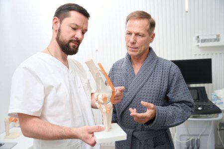 Foto de Qualified orthopaedist demonstrating a model of human knee joint to his patient - Imagen libre de derechos
