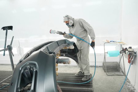 Téléchargez les photos : Man in protective suit standing in painting booth, holding spray gun and paint car bumper - en image libre de droit