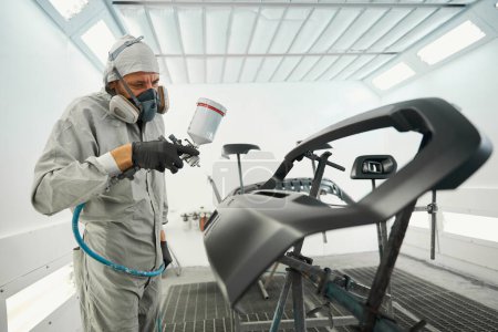 Professional man in protective suit standing in painting booth, holding spray gun and paint car details in tire fitting