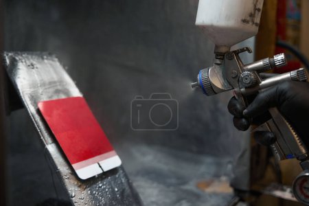 Photo for Man in protective gloves standing in painting booth, holding spray gun and paint testing card in red color - Royalty Free Image