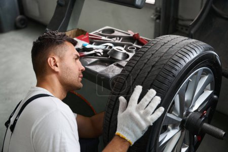 Téléchargez les photos : Mechanic in protective clothes and gloves standing near wheel and testing it in garage - en image libre de droit