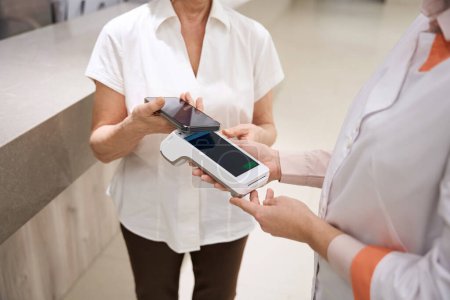 Photo for Patient pays for her visit to the therapist using the phone, the medical worker holds the terminal - Royalty Free Image