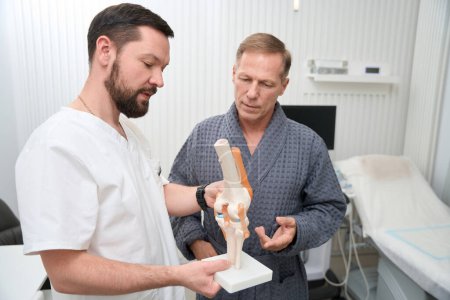 Foto de Professional orthopaedist holding a scientifically accurate model of knee joint while showing it to his patient - Imagen libre de derechos