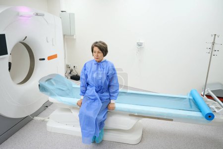 Foto de Elderly patient sits in the MRI diagnostic room on the moving part of the apparatus, the woman is saddened - Imagen libre de derechos