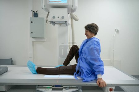 Foto de Optimistic elderly woman in special clothes is in the x-ray room, modern equipment is around - Imagen libre de derechos