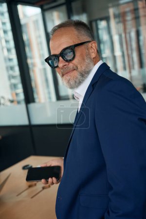 Téléchargez les photos : Waist-up photo of ambitious middle-aged businessman holding smartphone and smirking sneakily at the camera - en image libre de droit