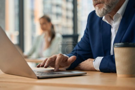 Téléchargez les photos : Cropped photo of older bearded man in business suit using his laptop at comfortable modern office shared with other coworkers - en image libre de droit