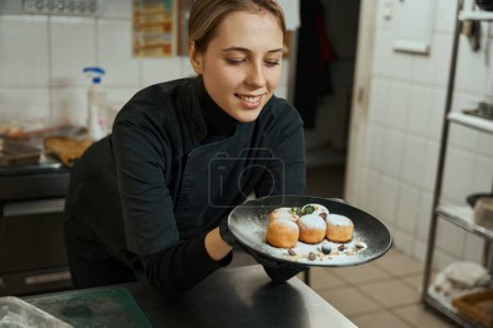Foto de Smiling woman looks at a plate with appetizing cheesecakes, the plate is decorated with berries and cream - Imagen libre de derechos