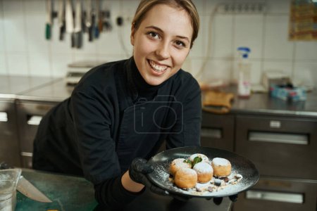 Foto de Cute chef holds in her hands plate with appetizing cheesecakes, the food is decorated with powdered sugar, berries and cream - Imagen libre de derechos