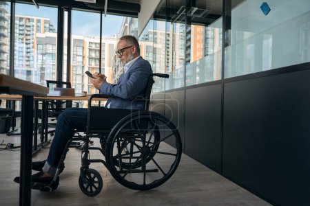 Téléchargez les photos : Homme d'affaires travaille dans un bureau dans un fauteuil roulant dans des vêtements de bureau occasionnels avec un téléphone dans ses mains - en image libre de droit