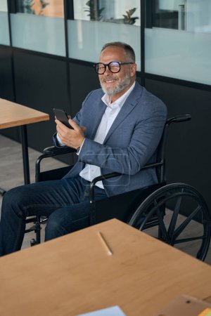 Téléchargez les photos : Optimiste homme d'âge moyen dans un fauteuil roulant avec un téléphone dans ses mains est au bureau - en image libre de droit