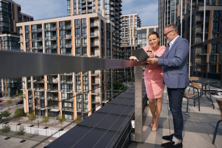 Téléchargez les photos : Businessman in business suit and glasses shows something in laptop to woman in beautiful dress, they stand against backdrop modern buildings - en image libre de droit