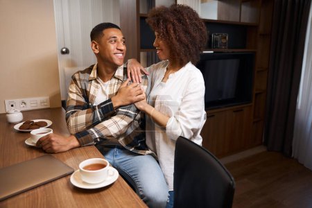 Téléchargez les photos : Charmant homme multiracial bavarder avec sa femme sur le café du matin dans la chambre d'hôtel, les gars en vêtements décontractés - en image libre de droit