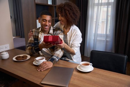 Téléchargez les photos : Smiling multiracial woman giving gift box to her boyfriend, couple having breakfast in hotel room - en image libre de droit