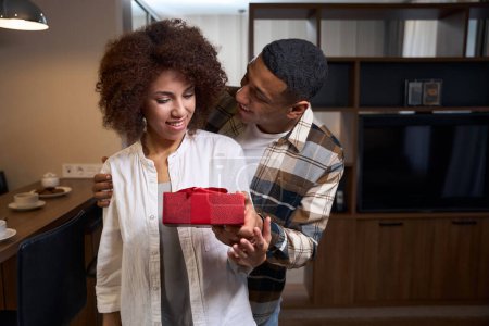 Foto de Happy multiracial guy gives his girlfriend a gift box, the couple is standing in the hotel room - Imagen libre de derechos