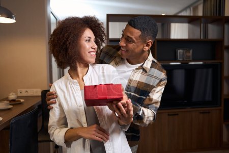 Foto de Chico feliz le da a su novia una caja de regalo roja, la pareja se mira con ternura - Imagen libre de derechos