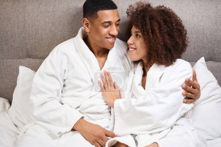 Téléchargez les photos : Curly woman and her boyfriend sit hugging on the bed, they look at each other with love - en image libre de droit