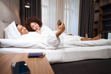 Téléchargez les photos : Charming multiracial couple comfortably settled on a bed in a hotel room, they are watching something on the phone together - en image libre de droit