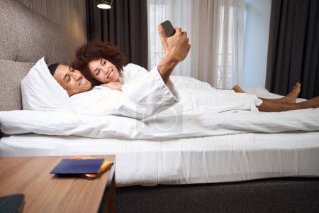 Foto de Viajar en pareja multirracial tomando selfie en la cama en la habitación del hotel, junto al pasaporte en la mesita de noche - Imagen libre de derechos