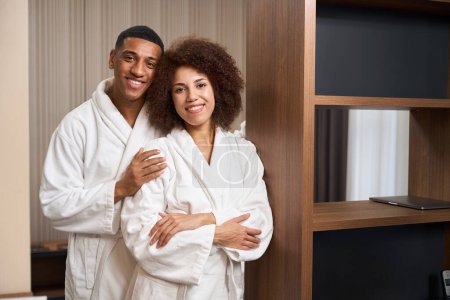 Photo for Multiracial smiling couple stands, embracing, in a hotel room, in a minimalistic design room - Royalty Free Image