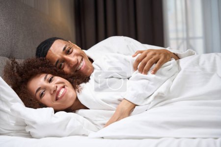 Téléchargez les photos : Souriant multiracial jeunes mariés couchés dans le lit dans la chambre d'hôtel, homme étreignant femme - en image libre de droit