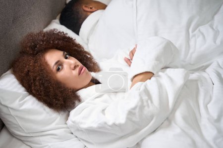 Photo for Frustrated young wife lies on the bed with her arms folded, next to her, turning away, her husband is sleeping - Royalty Free Image