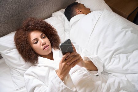 Téléchargez les photos : Jeune femme aux cheveux bouclés couchée au lit avec un téléphone, à côté d'elle, se retournant, son mari dort - en image libre de droit