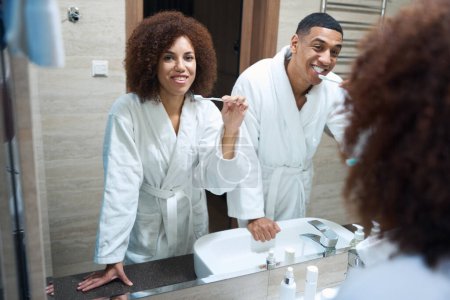 Foto de Husband and wife brush their teeth together in front of a mirror, it a beautiful multiracial couple - Imagen libre de derechos