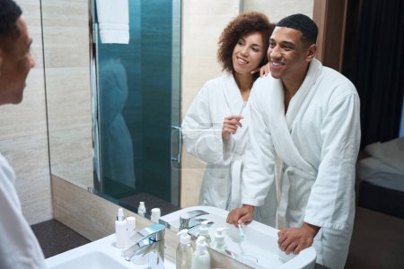 Foto de Multiracial hombre y su novia de pie con cepillos de dientes delante de un espejo, la pareja está vestida con albornoces - Imagen libre de derechos