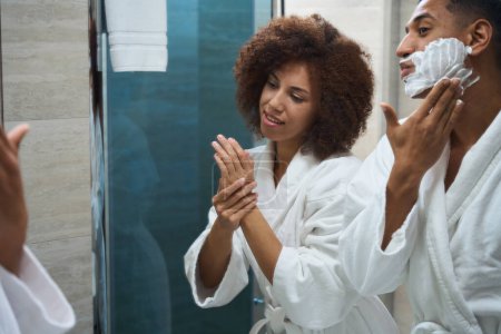 Téléchargez les photos : Jeune couple faisant la routine du matin dans la salle de bain de l'hôtel, homme appliquant mousse à raser pour faire face - en image libre de droit