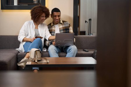 Téléchargez les photos : Multiracial couple sitting on sofa in front of laptop, woman pointing at screen - en image libre de droit