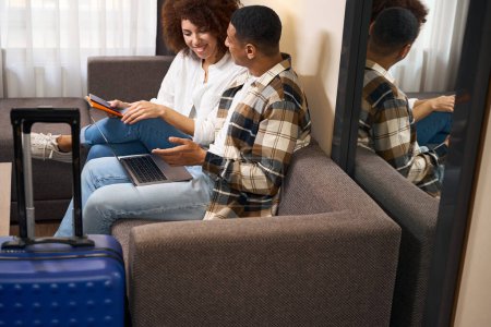 Téléchargez les photos : Les jeunes voyageurs sont assis sur un canapé dans une chambre d'hôtel avec des passeports et un ordinateur portable, les valises sont debout à proximité - en image libre de droit